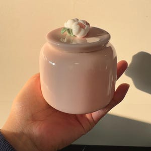 May include: A small, pink ceramic jar with a lid, topped with a white and pink flower. The jar is held in a hand, showcasing its smooth, glossy finish. The jar is a decorative item.