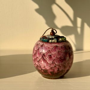 May include: A small, round, ceramic jar with a textured, scale-like pattern in shades of burgundy and pink. The lid is covered in a patterned fabric with a wooden bead and a brown string. The jar sits on a white surface.