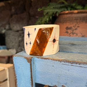 Puede incluir: Taza de cerámica hecha a mano con base crema, con una franja diagonal marrón salpicada de puntos negros. La taza está decorada con estrellas azules marino y un diseño de sol rojo y negro. La taza está sobre una superficie de madera azul desgastada.