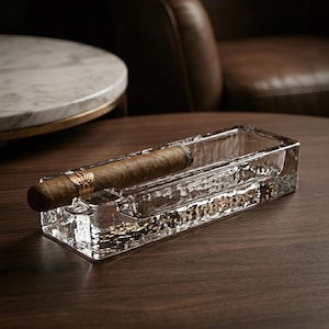 May include: A rectangular, textured glass ashtray with a lit cigar resting inside. The cigar is brown with a gold band. The ashtray sits on a dark wooden surface, with a blurred background of a table and leather chair.