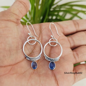 May include: A pair of silver earrings with a circular design and a small blue oval gemstone. The earrings have a hook closure and are displayed on an open hand. The earrings are a jewelry item.