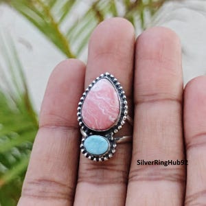 Puede incluir: Un anillo de plata con una piedra rosa grande en forma de pera con vetas blancas y una piedra azul claro ovalada más pequeña debajo. Ambas piedras están engastadas en plata con bordes de cuentas. El texto SilverRingHub92 es visible.