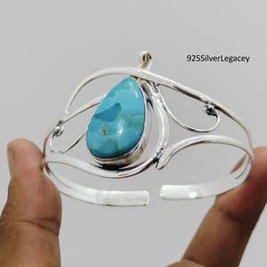May include: A silver cuff bracelet with a large, teardrop-shaped turquoise stone. The open-ended bracelet has a decorative, flowing design around the stone. The text "925SilverLegacy" is visible in the upper right corner.