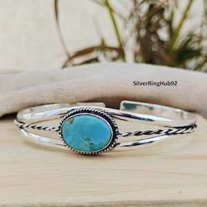 Könnte beinhalten: Ein silbernes Armreif-Armband mit einem großen ovalen Türkisstein in der Mitte. Der Türkis hat eine leuchtend blaue Farbe. Das Armband hat ein geflochtenes Seil-Design an den Seiten. Der Text "SilverRingHub92" ist sichtbar.