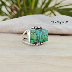 May include: A silver ring with a rectangular turquoise stone. The stone has a mix of green and blue hues with gold-colored veins. The ring has a double band design and decorative accents.