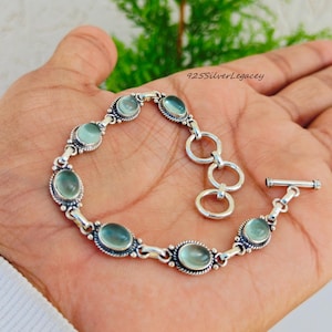 May include: A silver bracelet with oval, light blue gemstones. The bracelet is held in a hand, showcasing the design and the toggle clasp. The text "925SilverLegacy" is visible in the upper right corner.
