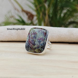 Op de afbeelding: Zilveren ring met een grote, rechthoekige steen. De steen heeft een gevlekt patroon met groen, rood en zwart. De ring ligt op een lichtgekleurd houten oppervlak. De tekst "SilverRingHub92" is zichtbaar.