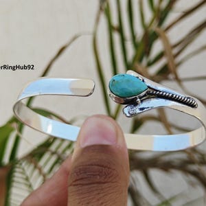 May include: A silver cuff bracelet featuring a turquoise teardrop-shaped stone. The open-design bracelet is held by a hand. The text "SilverRingHub92" is visible.