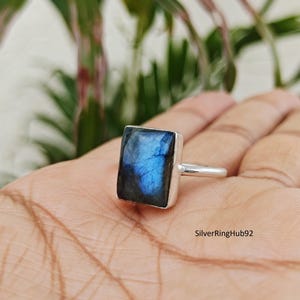 May include: A silver ring with a rectangular, iridescent blue gemstone. The gemstone displays shades of blue and black. The ring is set on a silver band. The ring is shown on a hand.