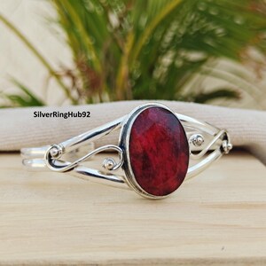Puede incluir: Una pulsera plateada con una gema ovalada de color rojo intenso en el centro. La pulsera presenta un diseño calado decorativo con detalles en espiral. El texto "SilverRinghub92" es visible en la imagen.