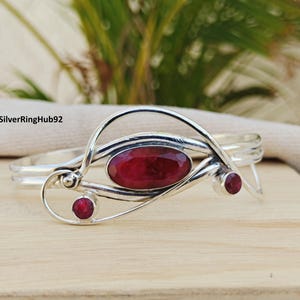 Könnte beinhalten: Ein silberfarbenes Armreif-Armband mit einem großen ovalen Rubin und zwei kleineren runden Rubinen. Das Armband hat ein offenes Design mit dekorativen Wirbeln und wird auf einer Holzoberfläche präsentiert.