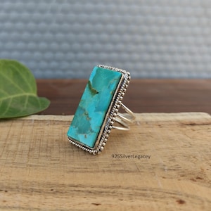 May include: A rectangular turquoise gemstone ring with a silver band. The turquoise stone is a vibrant blue-green color, set in a beaded silver frame. The ring has a multi-band design. The text "925SilverLegacy" is visible.