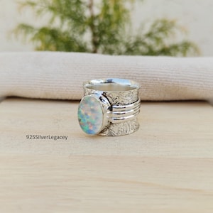 May include: A silver spinner ring featuring an oval-shaped iridescent opal gemstone. The band has an intricate floral-patterned texture and three horizontal silver bands. The text 925SilverLegacy is visible on the surface near the ring.
