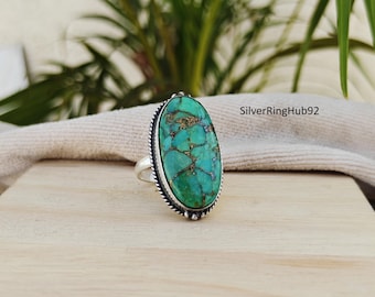 Puede incluir: Un anillo de plata con una piedra turquesa ovalada con patrones de matriz marrón. La piedra está engastada en un bisel de plata detallado con un borde de cuentas. El anillo se muestra sobre una superficie de madera.