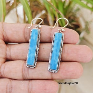May include: A pair of silver earrings with rectangular, light blue stones. The stones are set in a silver frame with a twisted rope design. The earrings have a simple hook closure. The earrings are displayed against a neutral background.