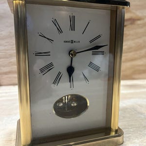 May include: A rectangular Howard Miller mantel clock with a polished gold-tone metal frame, a white face featuring black Roman numerals, and black hands. The clock includes a top carrying handle and a small oval window at the bottom.