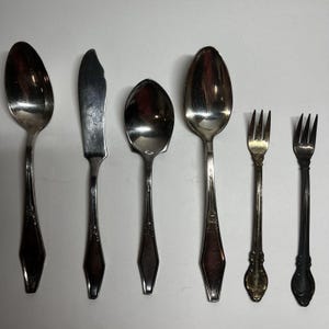 May include: A set of six silver-toned metal utensils including two large spoons, one butter knife, one medium spoon, and two small forks. Each piece features a polished finish and a tapered handle design with subtle decorative detailing near the neck.
