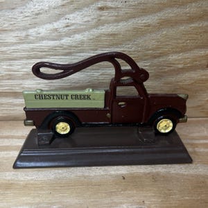 May include: A brown cast iron nutcracker shaped like a vintage pickup truck with a light green truck bed featuring the text CHESTNUT CREEK. The truck has yellow wheels and a long, curved handle extending from the roof to function as a lever.