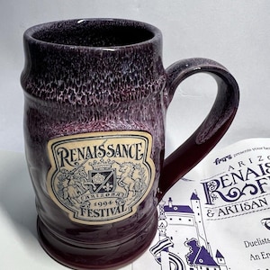 May include: A dark purple ceramic mug with a mottled glaze and a large handle. A cream-colored emblem on the front features the text "RENAISSANCE FESTIVAL" and "ARIZONA 1994" with a central shield, a crown, and two unicorn figures.