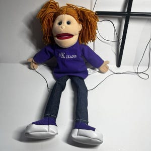 May include: A marionette puppet with orange yarn hair, a beige face, and a purple long-sleeved shirt featuring the text OK JEANS in white embroidery. The puppet wears dark blue denim pants and purple shoes with white soles, controlled by black strings.