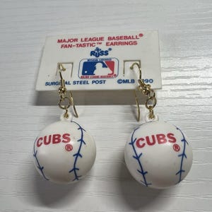 May include: A pair of dangling earrings featuring miniature white baseballs with red CUBS text and blue stitching. The earrings are attached to a white display card labeled MAJOR LEAGUE BASEBALL FAN-TASTIC EARRINGS and RUSS OFFICIAL LICENSEE.