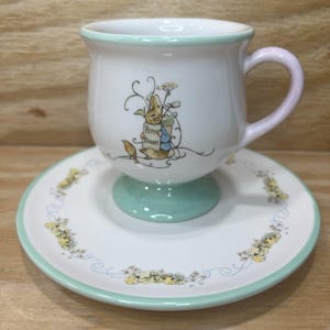 May include: A white ceramic teacup and saucer set. The cup has a light green base and a pink handle. The saucer and cup are decorated with a floral design and a Peter Rabbit illustration. The cup reads "Peter Rabbit".