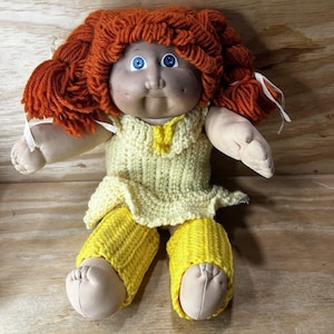 May include: A doll with bright orange yarn hair styled in pigtails, blue eyes, and a round face. The doll wears a pale yellow crocheted dress with a matching yellow collar and bright yellow crocheted leg warmers over its fabric legs and feet.