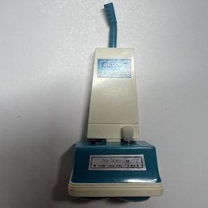 May include: A vintage Galoob Fun Beam toy vacuum cleaner featuring a cream-colored upright body with a blue handle and a teal base. The front of the unit displays a silver label with the text Fun Beam and a series of decorative dots and dashes.