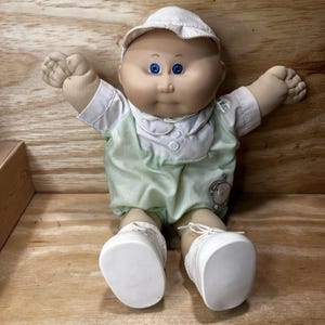 May include: A doll with a round face, blue eyes, and a bald head wearing a white cap. The doll has a soft fabric body, wearing a white collared shirt with two buttons, a light green romper with an elephant graphic, and white lace-up shoes.