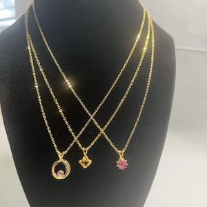 May include: Three gold-toned necklaces with delicate chains. The left pendant is a textured gold circle with a small pink stone. The center pendant is a faceted amber-colored stone. The right pendant is a faceted pink stone in a gold setting.