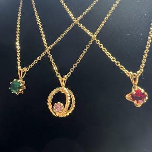 May include: Three gold-tone necklaces with delicate chains. The left pendant features a round green gemstone, the center pendant is a circular frame with a small pink stone, and the right pendant holds a round red gemstone in a prong setting.