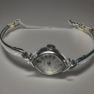 May include: A silver-tone Caravelle wristwatch featuring an oval-shaped case, a white dial with silver markers, and a numeral 12. The watch includes a flexible metal expansion band with gold-tone accents and a thin safety chain for secure closure.