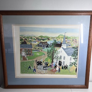 May include: A framed folk art print depicting a rural village scene with a white church, a horse-drawn carriage, and various figures in a landscape. The artwork features a light blue matting and a brown wooden frame, capturing a detailed community gathering.