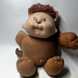May include: A brown plush cat doll with a smooth, molded vinyl face, dark brown yarn hair, and small, round, dark eyes. The body is made of soft brown fabric with stitched paw details on the feet and hands, and a small, round belly button feature.