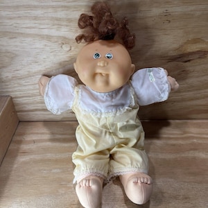 May include: A vintage Cabbage Patch Kid doll with a round face, blue eyes, and curly brown hair. The doll is wearing a yellow jumpsuit with white lace trim and a white bib. The doll's arms are outstretched.