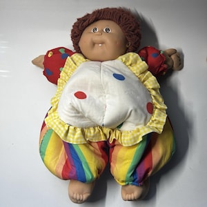 May include: A soft-sculpture doll with brown yarn hair, a round face, and a single visible tooth. The doll wears a white bib with red and blue polka dots, yellow gingham ruffles, red sleeves with colorful hearts, and rainbow-striped pants.