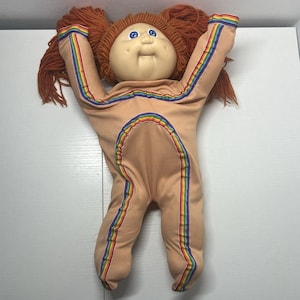 May include: A soft-bodied doll with a round vinyl head, blue eyes, and orange yarn hair styled in two pigtails. The doll wears a tan one-piece outfit featuring rainbow-colored stripes running down the arms, legs, and in a curved arch across the torso.