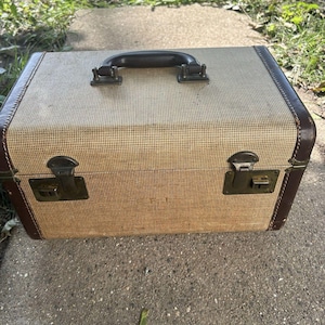 May include: A rectangular hard-shell travel case featuring a textured beige fabric exterior with dark brown leather-like trim. The case includes a dark brown top handle, two metal latches with keyholes on the front, and reinforced corner edges.
