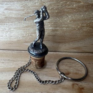 May include: A silver-toned bottle stopper featuring a golfer in mid-swing. The stopper is attached to a chain and a ring. The cork base is visible. The overall design is detailed and decorative.