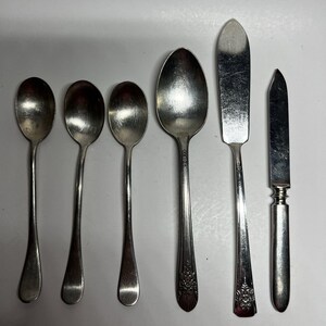 May include: A set of six silver-toned metal utensils arranged in a row, including three small spoons with plain handles, one large spoon with a floral pattern on the handle, one flat-edged spreader, and one narrow knife with a decorative handle base.