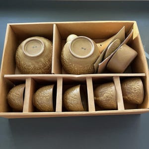 May include: A tea set in a cardboard box, featuring a teapot and six matching cups. The ceramic pieces are a muted tan color with a textured, embossed floral pattern. Each piece has a small, dark character mark on the bottom surface.