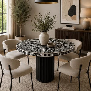 May include: A round dining table with a black and white patterned top and a black pedestal base. Four beige chairs with black legs surround the table. A vase with greenery sits on the table, and a black pendant light hangs above.