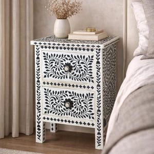 Może przedstawiać: Biała szafka nocna z dwiema szufladami, ozdobiona czarnymi wzorami kwiatowymi. Na górze wazon z suszonymi kwiatami i książki. Boki szafki mają dekoracyjny wzór. Stoi obok łóżka.