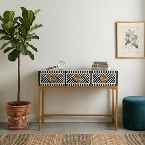May include: A console table with a black and white geometric design on the drawers and gold handles. The table has a gold metal frame. A potted plant, a framed botanical print, and a teal ottoman are also in the room. The table measures approximately 90 cm in width.