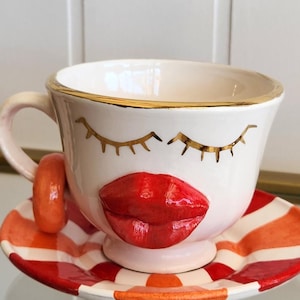 May include: A white ceramic teacup and saucer set with a whimsical design. The cup features a red lip design and gold eyelashes. The saucer has red and orange stripes. The cup has a gold rim.