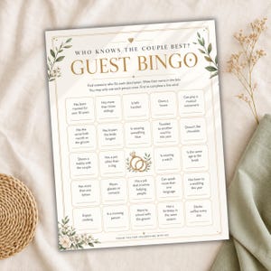 May include: A rectangular wedding guest bingo game card with gold text reading Who Knows The Couple Best? Guest Bingo. The card features a grid of twenty-five squares with prompts, a central illustration of two gold rings, and green leaf accents.