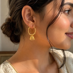 Könnte beinhalten: Ein goldener Creolen-Ohrring mit einem kleinen, glockenförmigen Jhumka-Anhänger. Der Reifen ist entlang seiner unteren Kurve mit einer Reihe winziger, texturierter Goldperlen verziert, während der zentrale glockenförmige Anhänger filigrane Metallarbeiten zeigt.