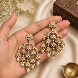 May include: A pair of gold-toned chandelier earrings featuring intricate filigree metalwork, encrusted with numerous clear white crystals, and finished with small, dangling white pearls at the base of each tiered, bell-shaped component.