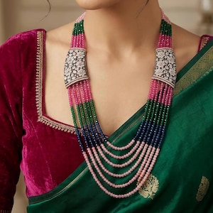 Può includere: Una collana a più fili con perline rosa, verdi e blu, accentuata da chiusure argentate ornate. Sono visibili anche orecchini abbinati con una pietra rossa pendente. I gioielli sono ambientati su uno sfondo di un top di velluto rosso scuro e un indumento di seta verde.