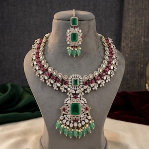 Puede incluir: Un lujoso conjunto de collar y tocado. El collar presenta un colgante central de piedra verde talla esmeralda, rodeado de piedras blancas y rojas más pequeñas. El tocado a juego tiene un diseño similar, con una sola piedra verde talla esmeralda y cuentas verdes colgantes.
